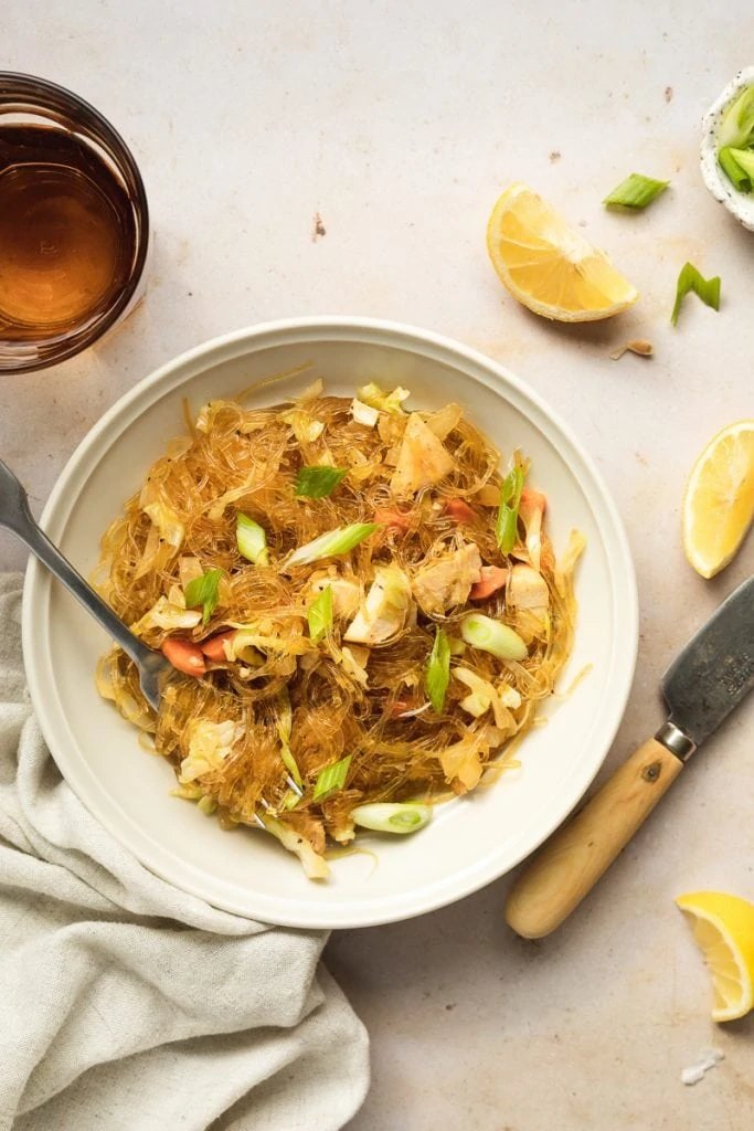 Filipino pancit in a bowl with lemon wedges and a brown glass