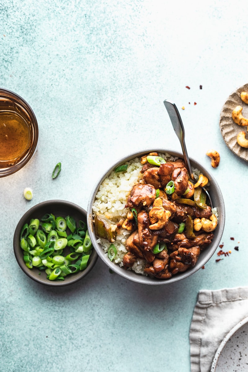 Keto cashew chicken in a bowl on a teal backdrop with a glass of water. 