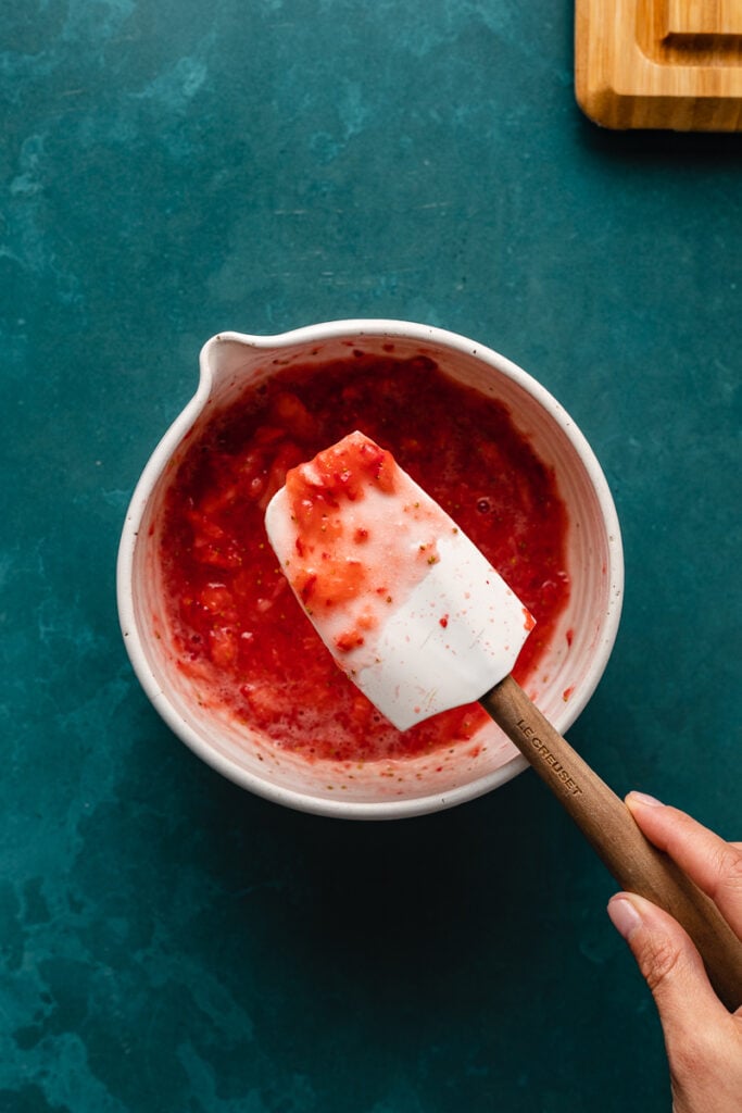White rubber spatula held over a bowl of fresh strawberry mixture for keto strawberry mousse recipe.
