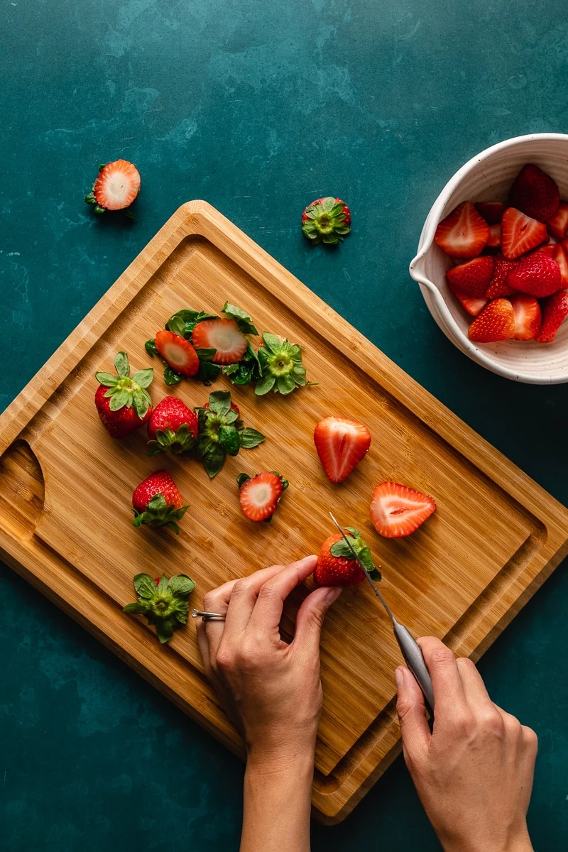 Cutting fresh strawberries on a wood cutting board on a turquoise counter.