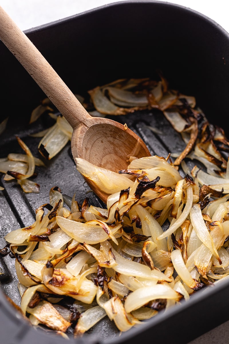 Air fryer onions being scooped up with a wooden spoon in air fryer basket.