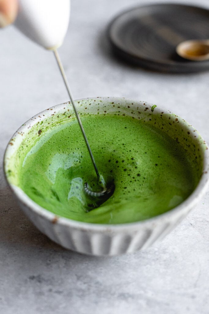 Frothing matcha powder with warm water in a speckled dish.