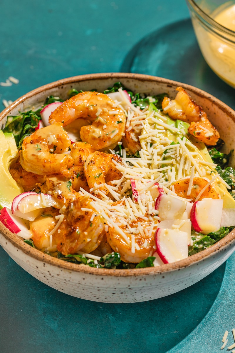 A bowl of kale caesar cajun shrimp salad with dressing behind it.