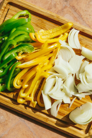Green bell pepper, yellow pepper and onions sliced on a cutting board.
