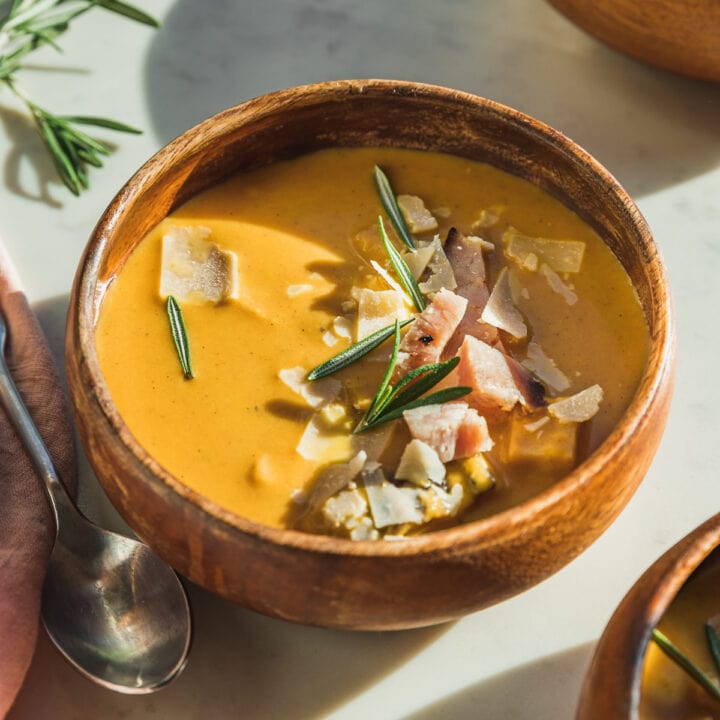 A bowl of butternut squash and carrot soup with chopped ham, parmesan and rosemary on top.