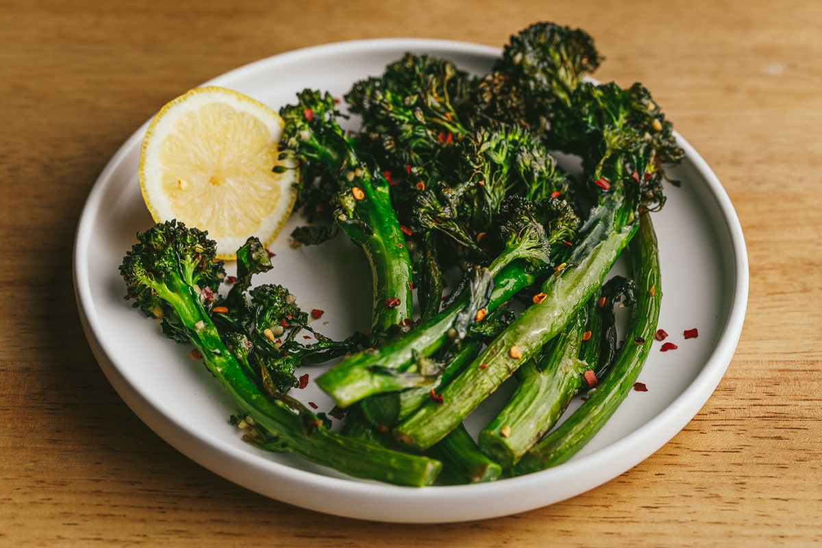 Air fryer broccolini on a white plate with red pepper flakes and a slice of fresh lemon.