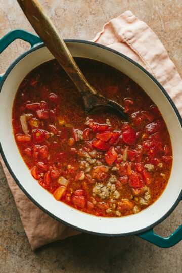 Diced tomatoes, beef and aromatics in a large pot with a wooden spoon.
