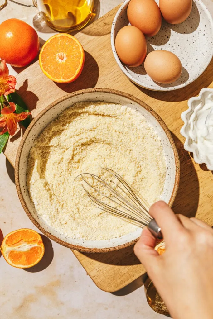 Stirring dry ingredients in a mixing bowl with oranges and eggs on the side.