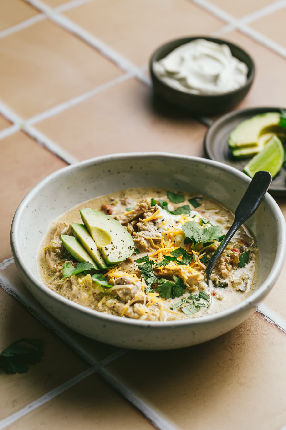 Crockpot green chili chicken soup in a serving bowl with toppings on a beige tile surface.