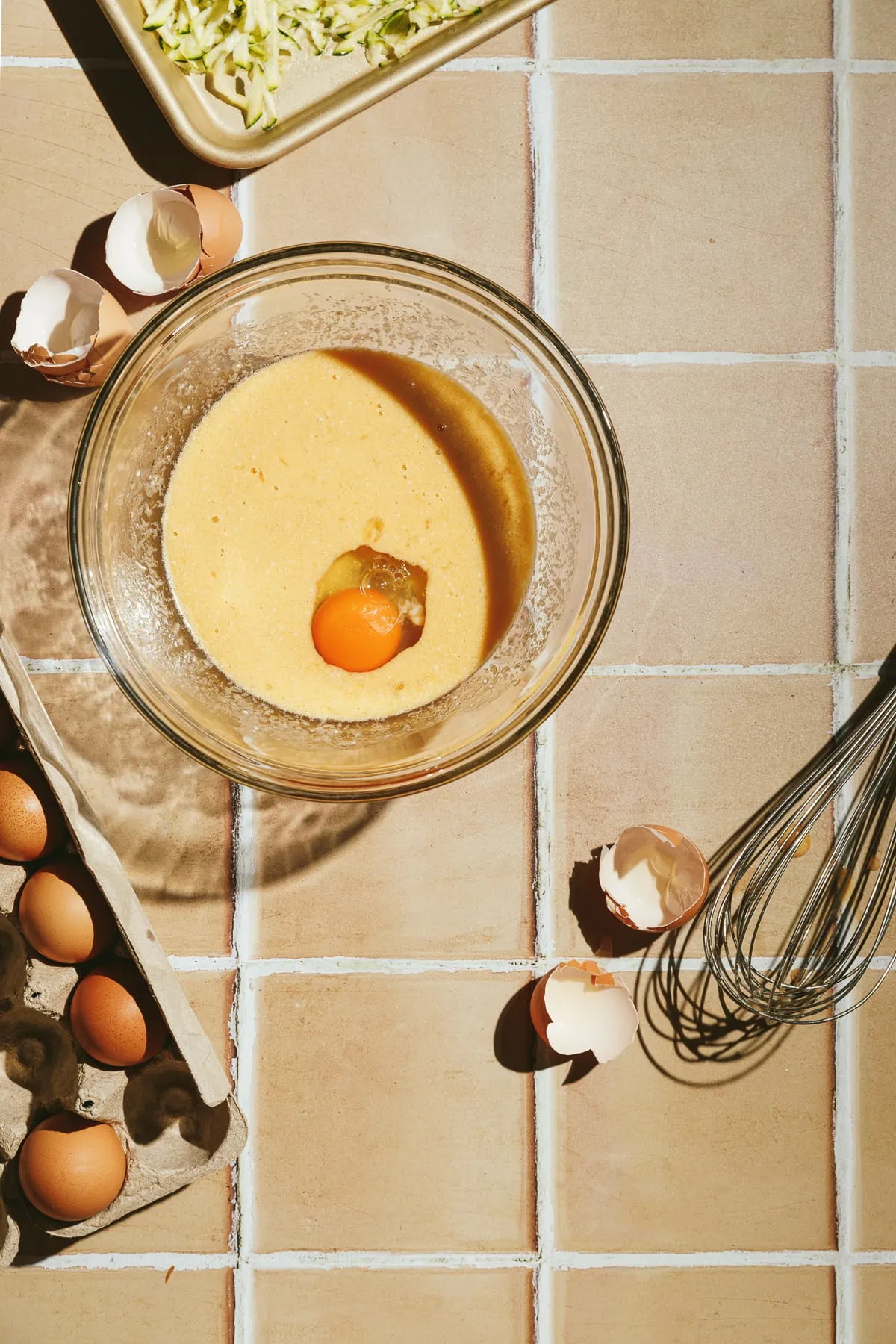 Mixing bowl, cracked eggs and a whisk on a tile surface.