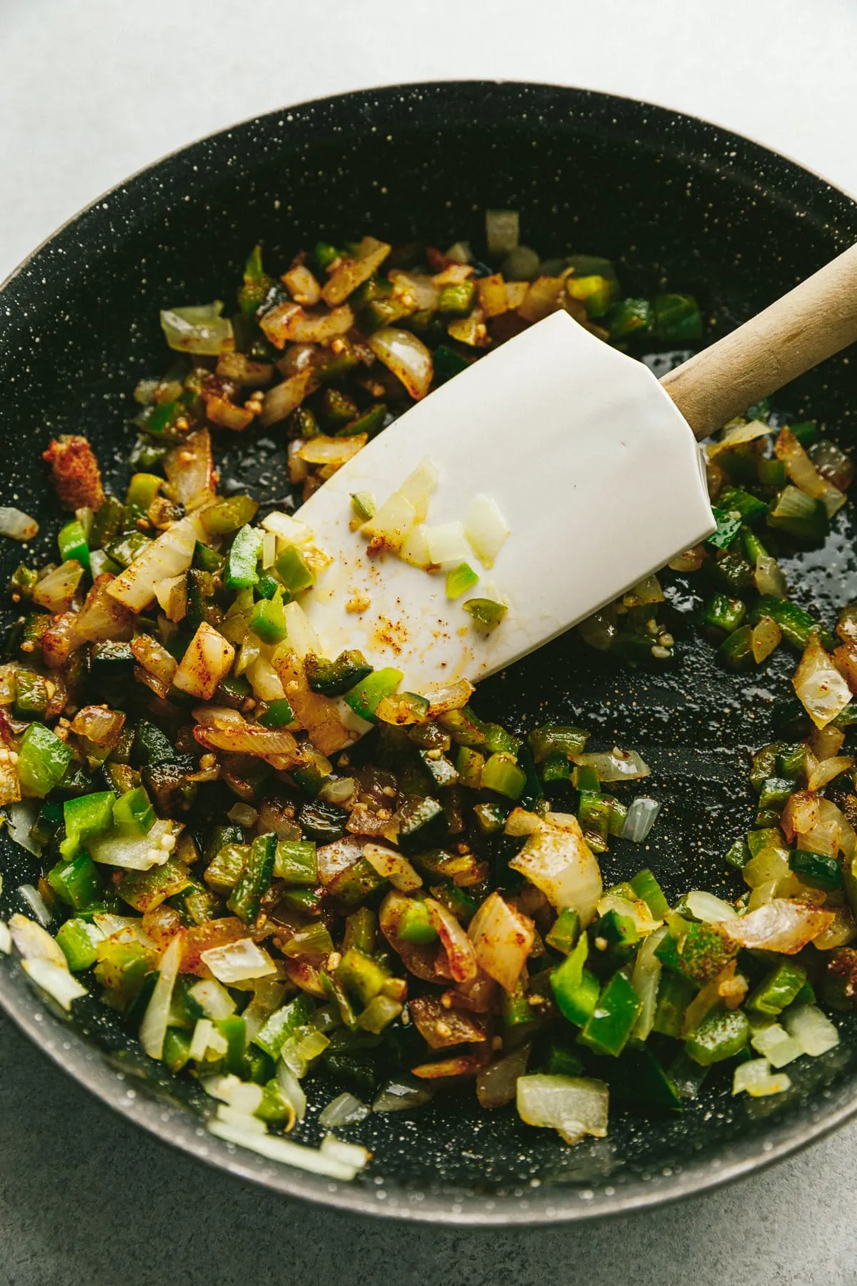 A spatula stirring chopped green chilli peppers and onions in a pan.