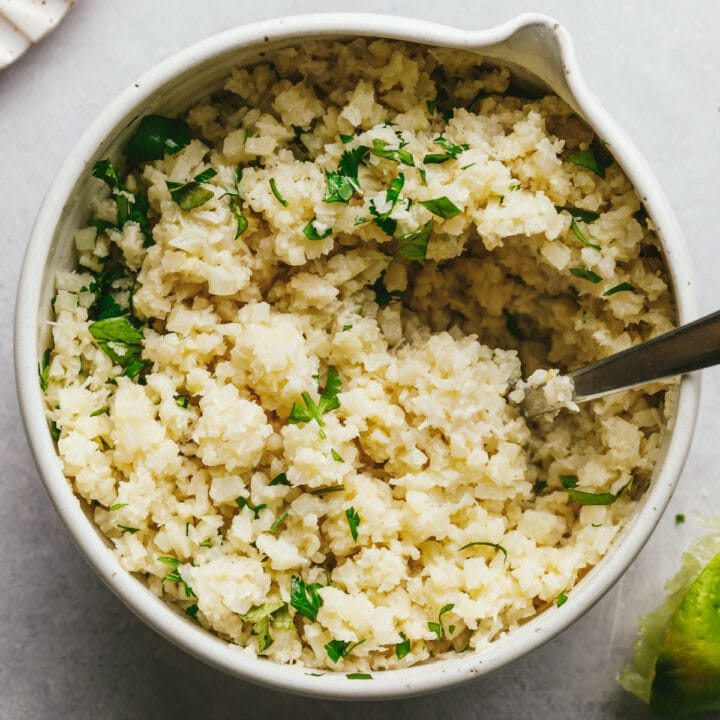 Coconut cauliflower rice in a white bowl with a serving spoon and garnish of fresh cilantro.