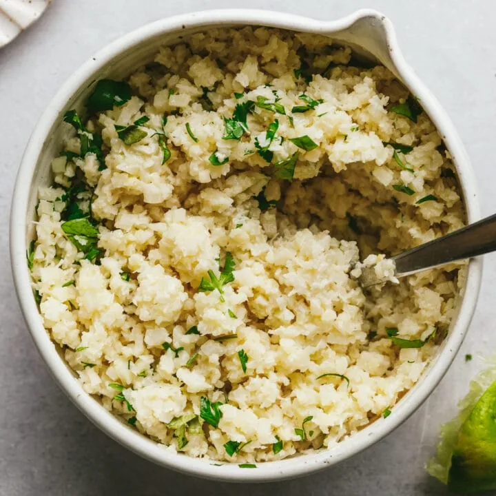 Coconut cauliflower rice in a white bowl with a serving spoon and garnish of fresh cilantro.