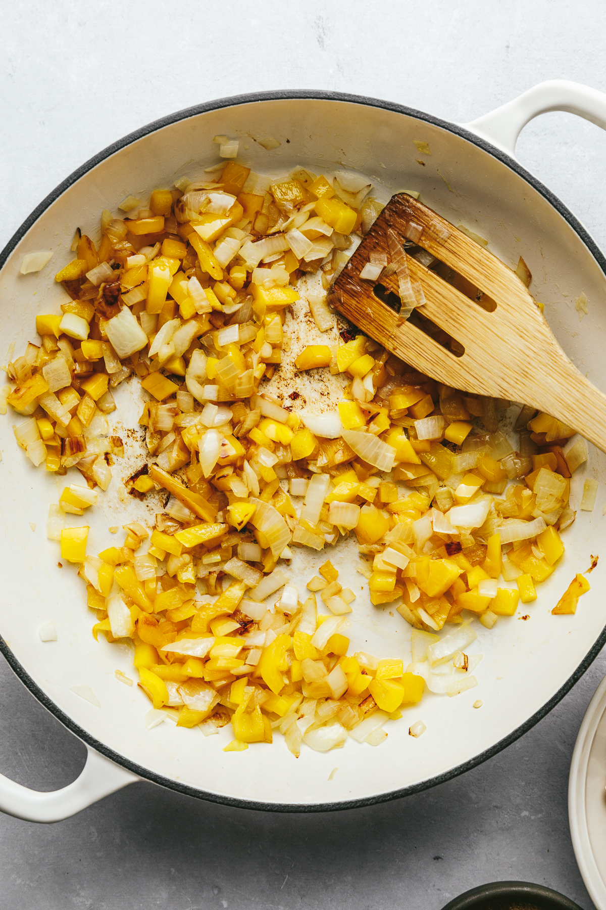 Diced onions and yellow bell pepper in a white skillet with a wooden spatula.