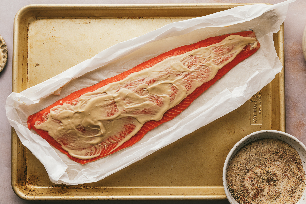 A filet of salmon in parchment paper on a baking sheet with Dijon mustard rubbed on top.