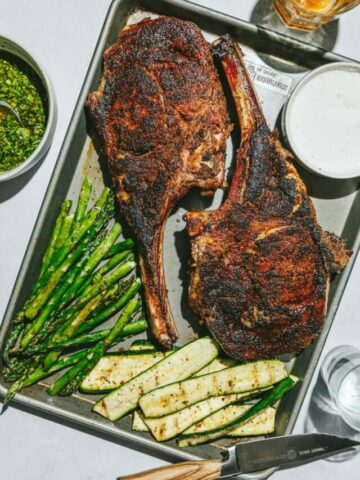 A baking sheet with two tomahawk steaks and grilled vegetables.