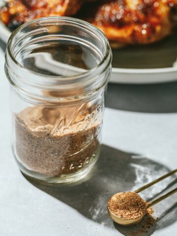 Keto bbq rub in a jar with a teaspoon next to it and grilled chicken in the background.