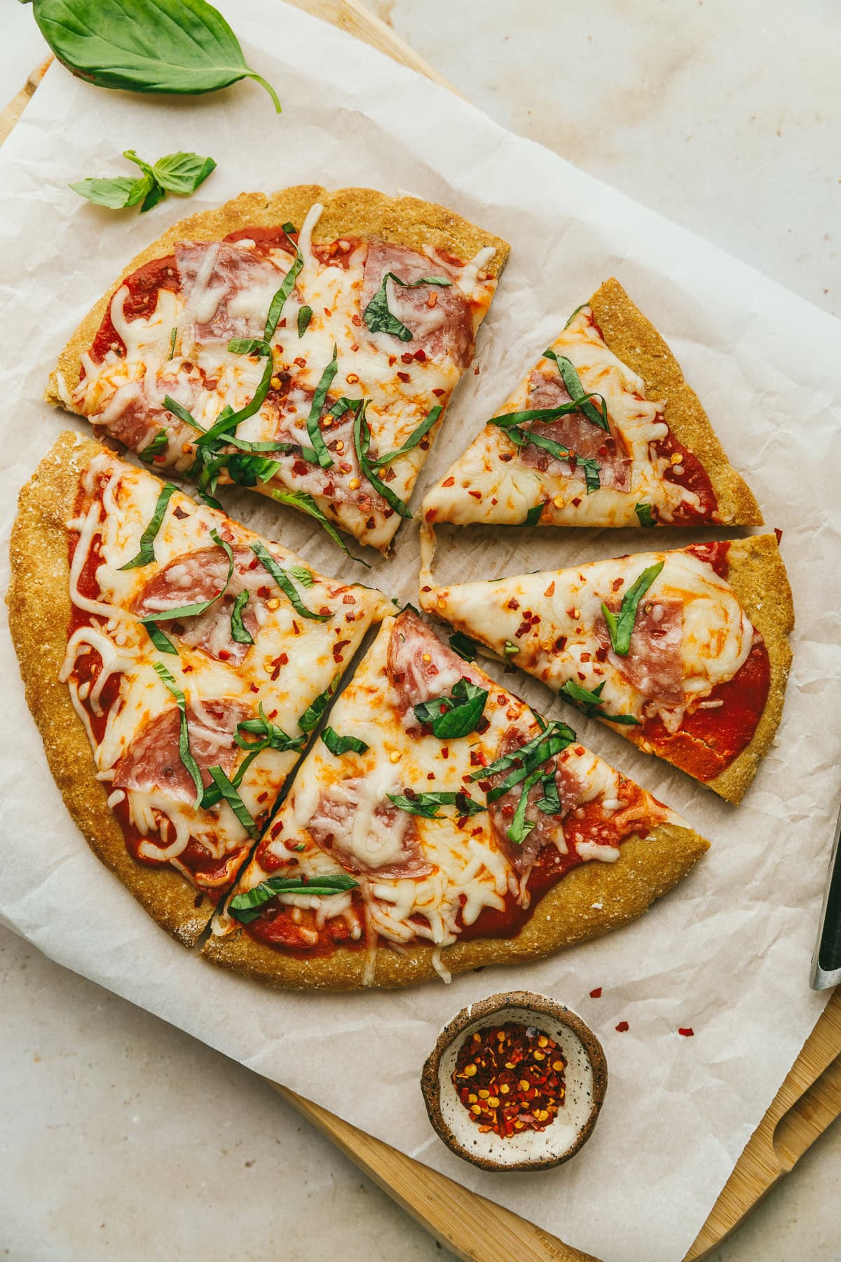 A grain-free pizza on white parchment paper with fresh basil and red pepper flakes.